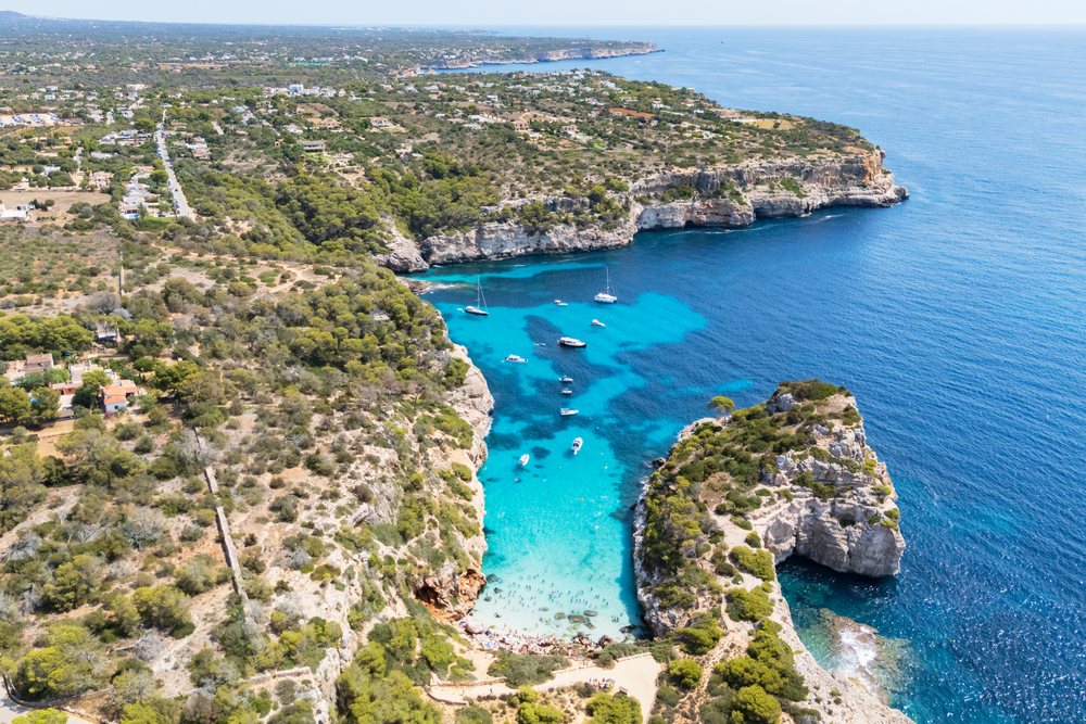 Aerial view of Caló del Moro cove with clear blue water, boats anchored, beach, and surrounding cliffs and forest, Landscape, Lifestyle, Mallorca, Spain.