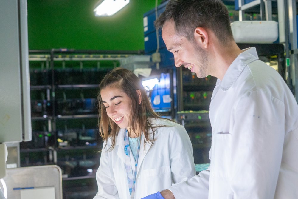 Two smiling scientists wearing lab coats collaborate on research in a laboratory setting, Spain.