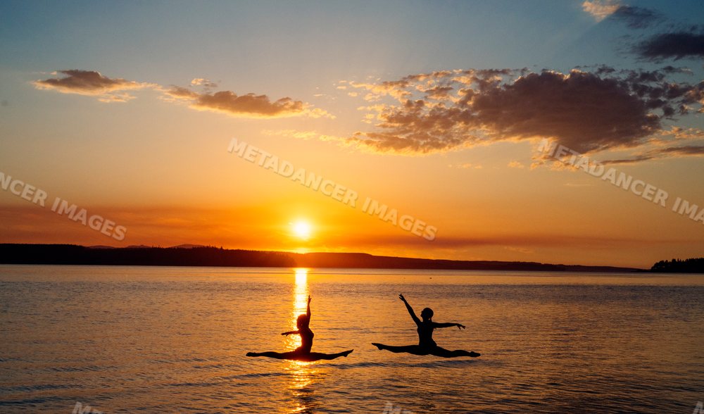 Two white people performing jumping split poses at sunset on a lake