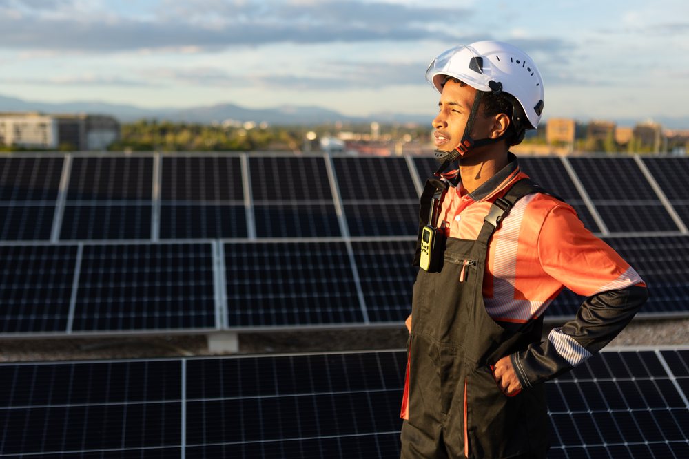 Solar energy technician wearing a white safety helmet and orange-black overalls stands outdoors amid solar panels at a solar farm, reflecting sunlight, Madrid, Spain.
