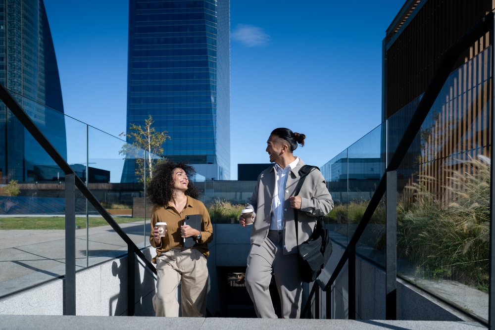 Two adults walking up outdoor stairs holding coffee cups and chatting in an urban area on a sunny day, Madrid, Spain.