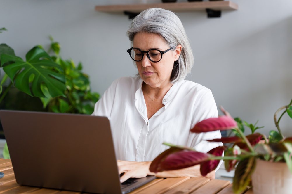 Woman with gray hair and glasses typing on a laptop at a wooden table surrounded by plants in a home office, Spain.
