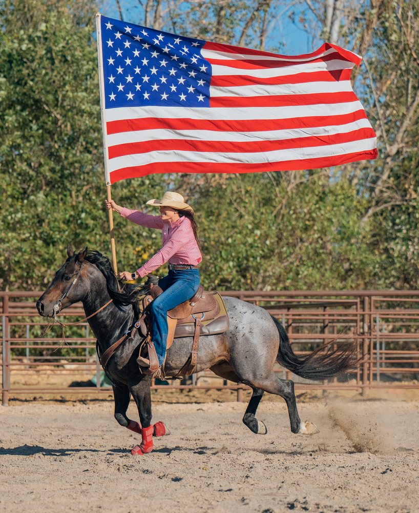 Woman wearing a western hat rides a galloping horse while holding an American flag in a rodeo arena, Southwest USA.