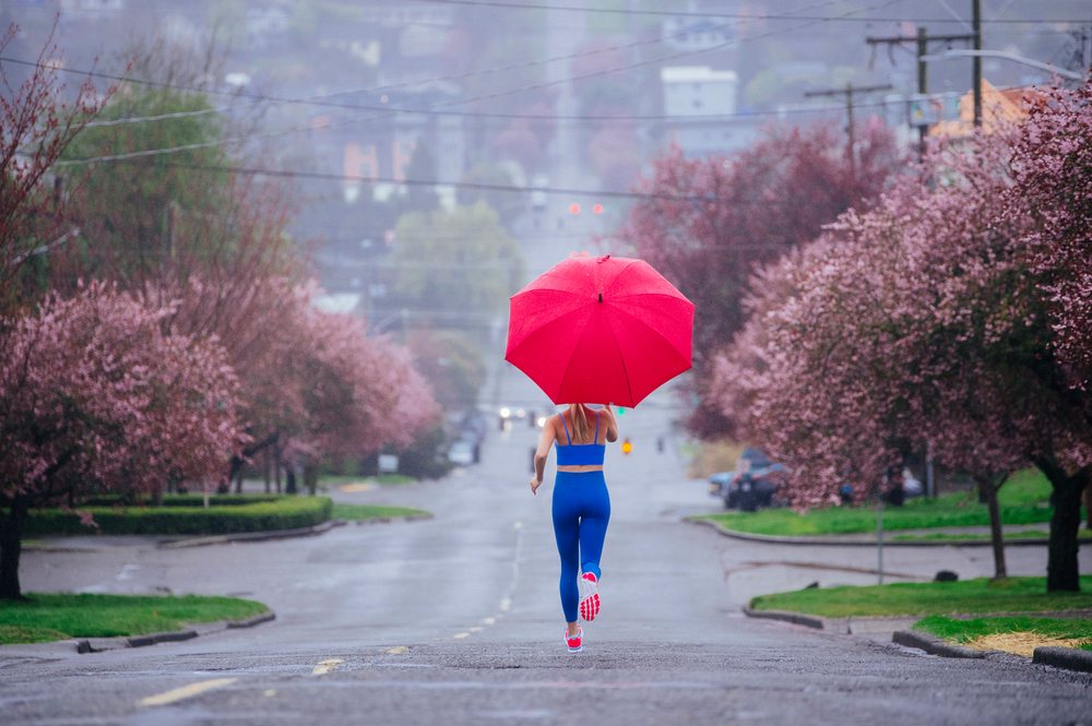 Woman wearing blue sportswear runs with a red umbrella on a rainy street lined with pink flowering trees in an urban area of the, USA.