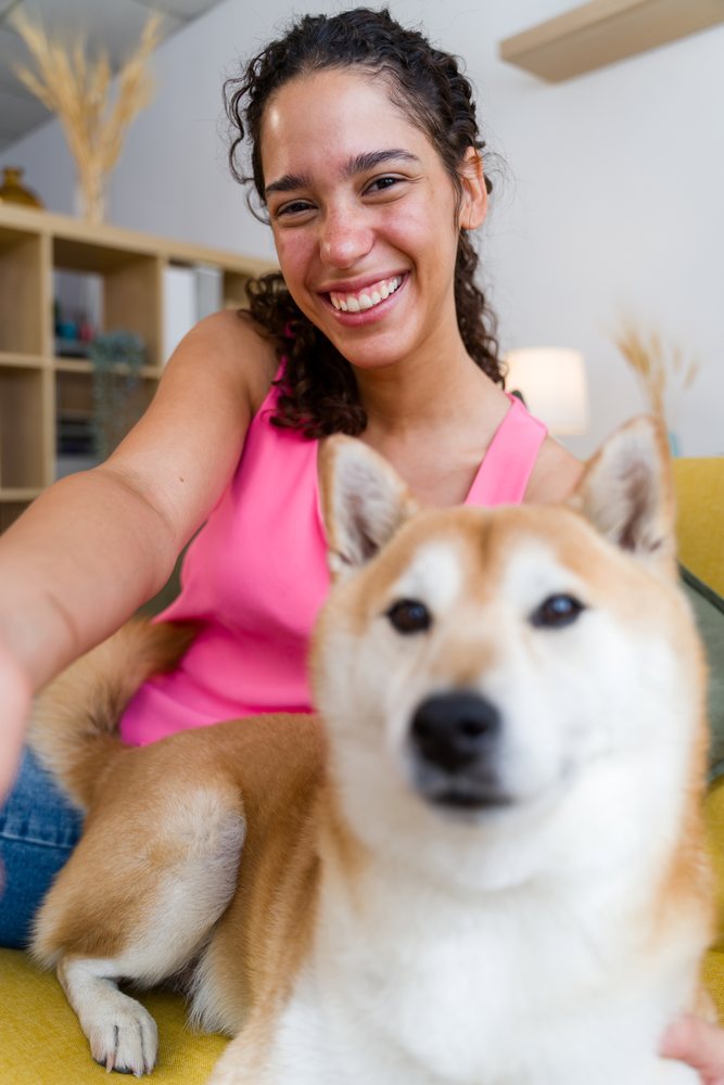 A woman in a pink shirt smiles happily as she takes a selfie with her small dog on a yellow sofa in a living room, Spain.