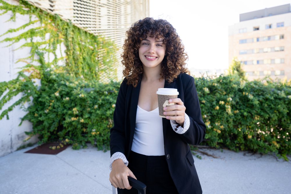 Woman with curly hair wearing a black blazer and white top holds a coffee cup smiling outdoors in an urban area with green foliage, Spain.