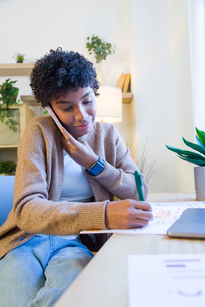Woman talking on phone and writing in a notebook at a home office desk with a laptop and indoor plants, remote work.