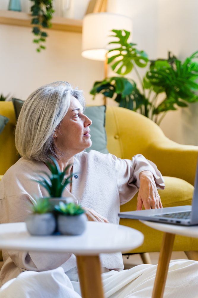 Woman with grey hair sitting on floor leaning on yellow sofa beside laptop and white table with potted plants indoors, Spain.