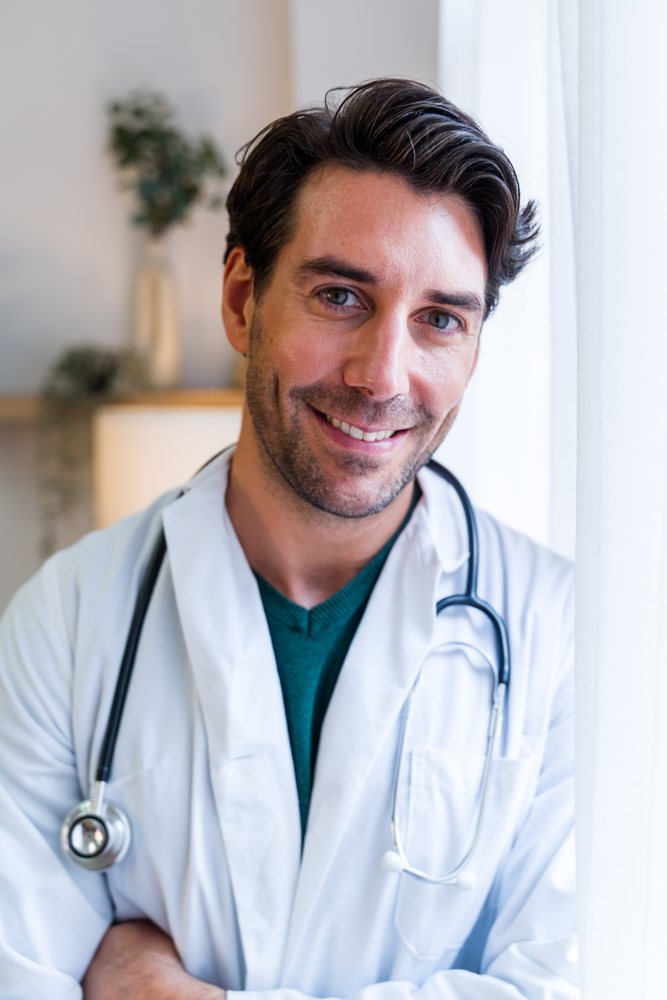 Male adult doctor wearing a white coat and stethoscope smiles indoors near a window in a clinical setting with plants visible in background, Spain.