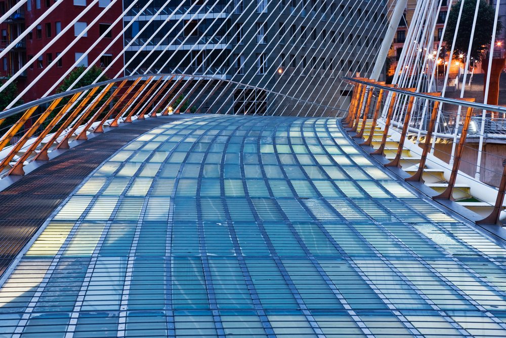Zubizuri pedestrian bridge with curved glass floor and steel cables illuminated in the evening, Bilbao, Spain.