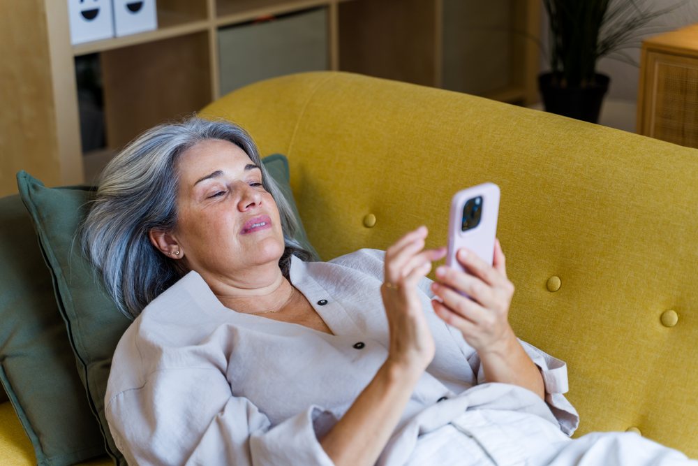 Middle-aged woman with gray hair holding a smartphone while relaxing on a yellow couch indoors, Spain.