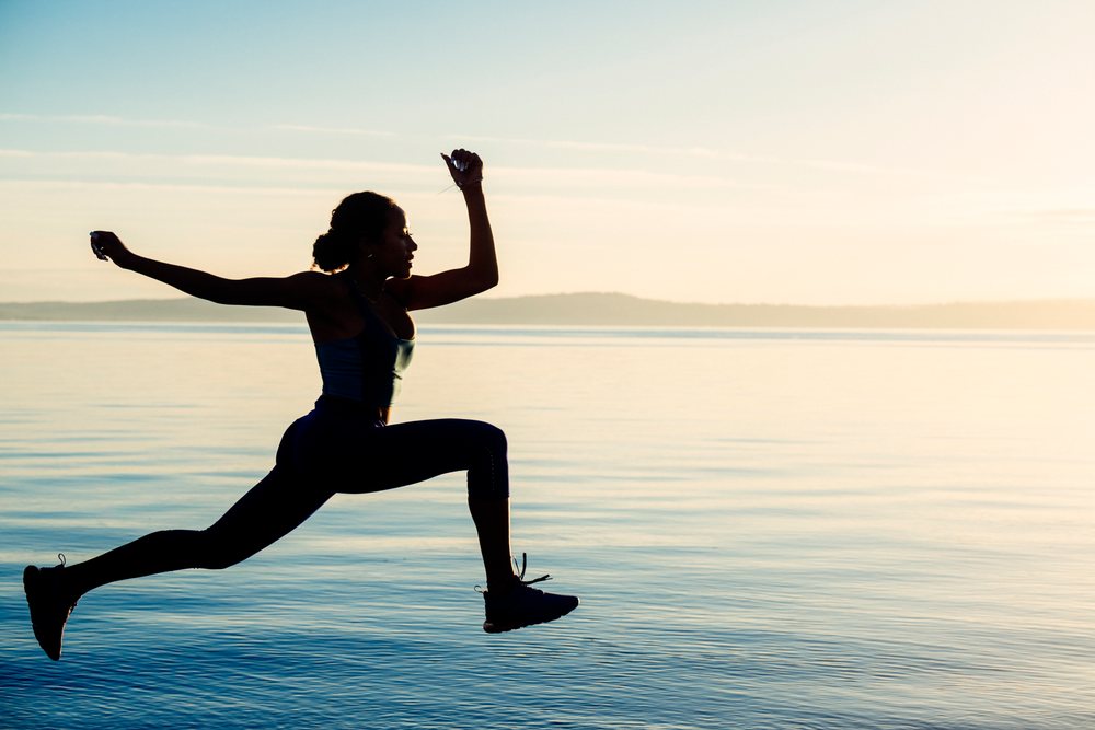 Woman jumping in silhouette exercise pose outdoors at sunset by a lake, yoga, acrobatics, USA.