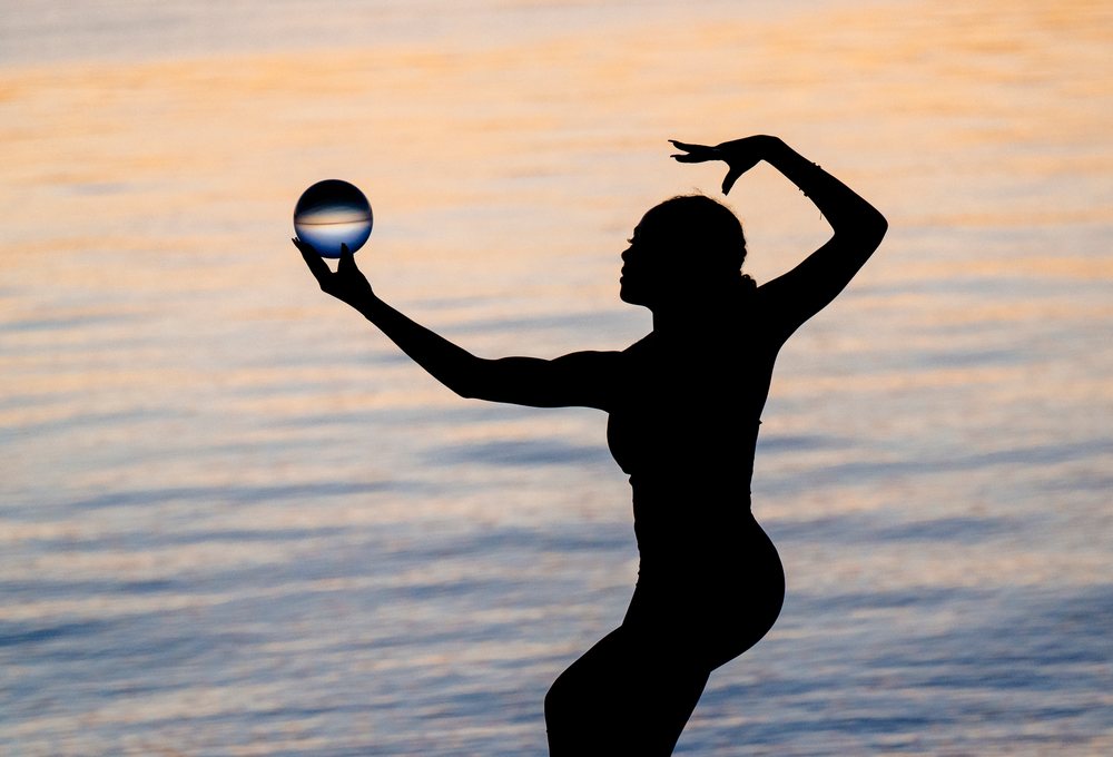 Silhouette of a woman holding a glass ball and performing a yoga acrobatic balance pose by the water at sunset, USA.