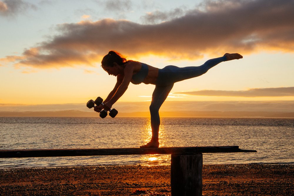 Woman balancing in a yoga pose while holding dumbbells at sunset on a beach with water in the background, WA, USA.