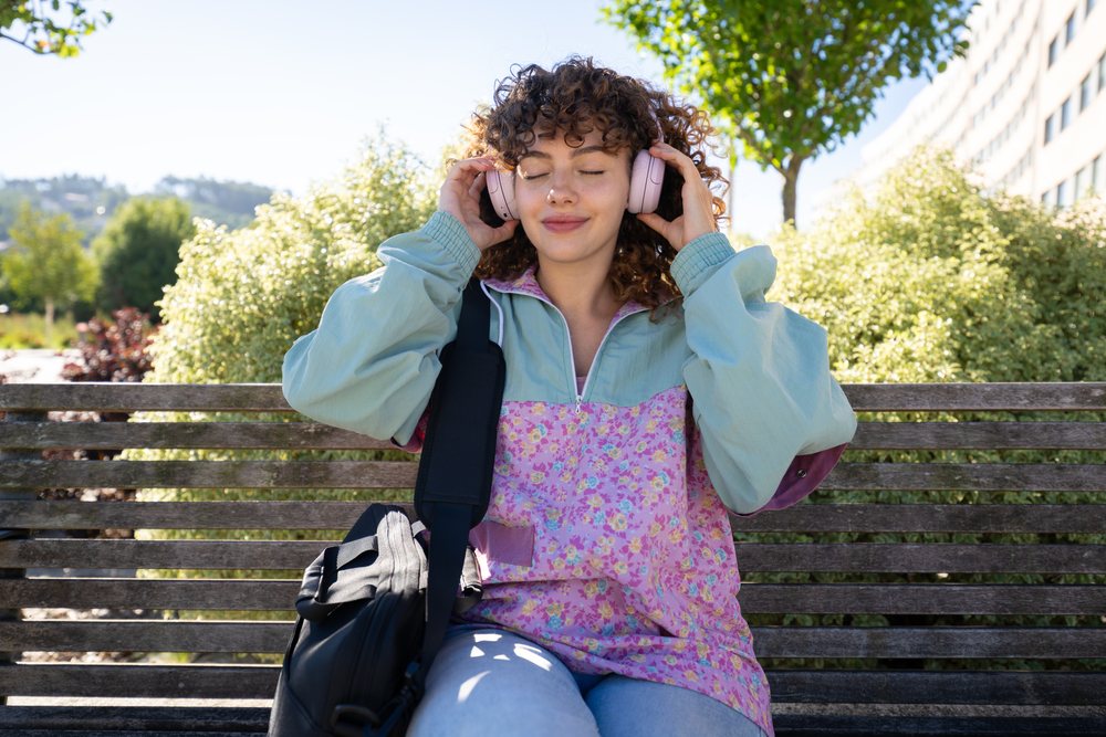 Woman with headphones sitting on a wooden bench in a sunny park, Spain.