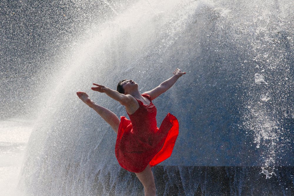Female dancer in red dress leaps gracefully through water spray outdoors, USA.