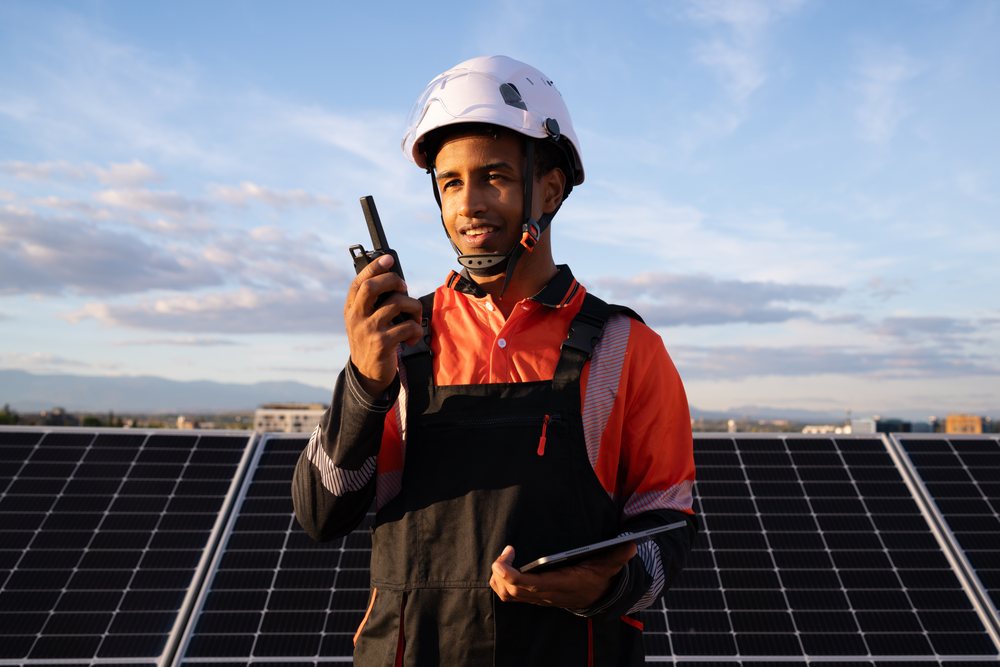Technician wearing safety helmet communicates with walkie talkie and inspects solar panels outdoors at sunset, Madrid, Spain.