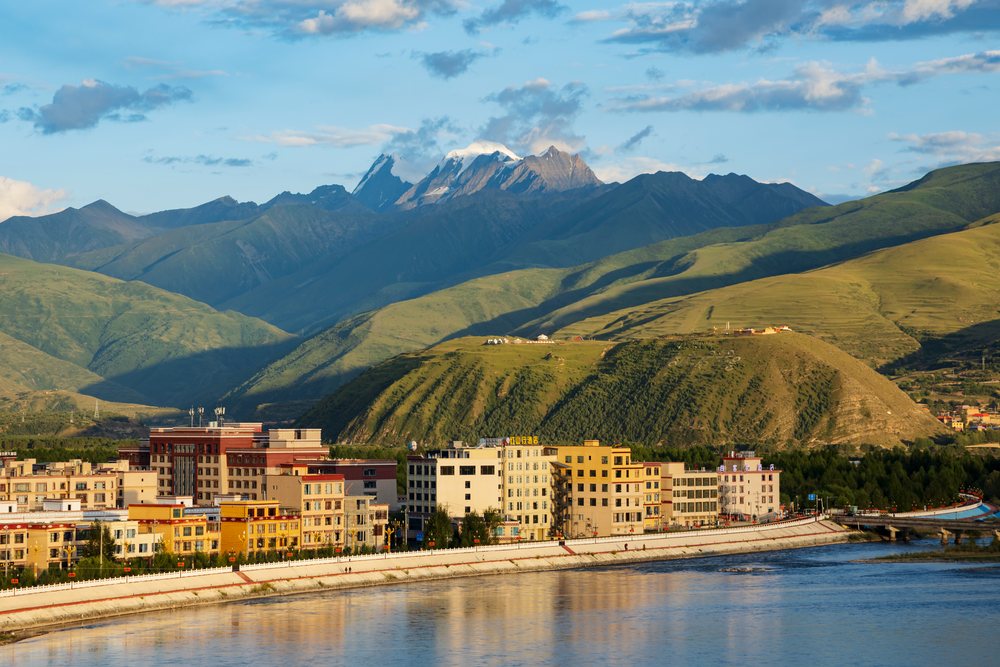 Sunset view of the Ganze River with reflective water, urban buildings, green hills, and snow-capped Kawaluori Mountain, China, Sichuan Province, People's Republic of China.