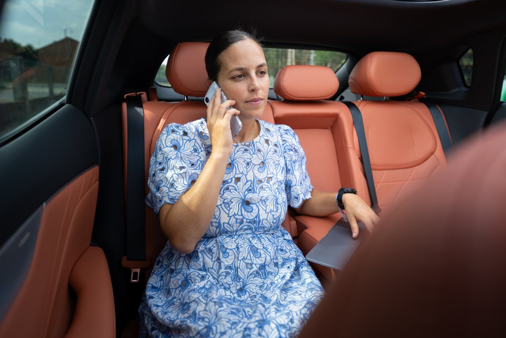 Woman sitting in the back seat of a car, talking on her phone while holding a silver laptop on her lap, Spain.