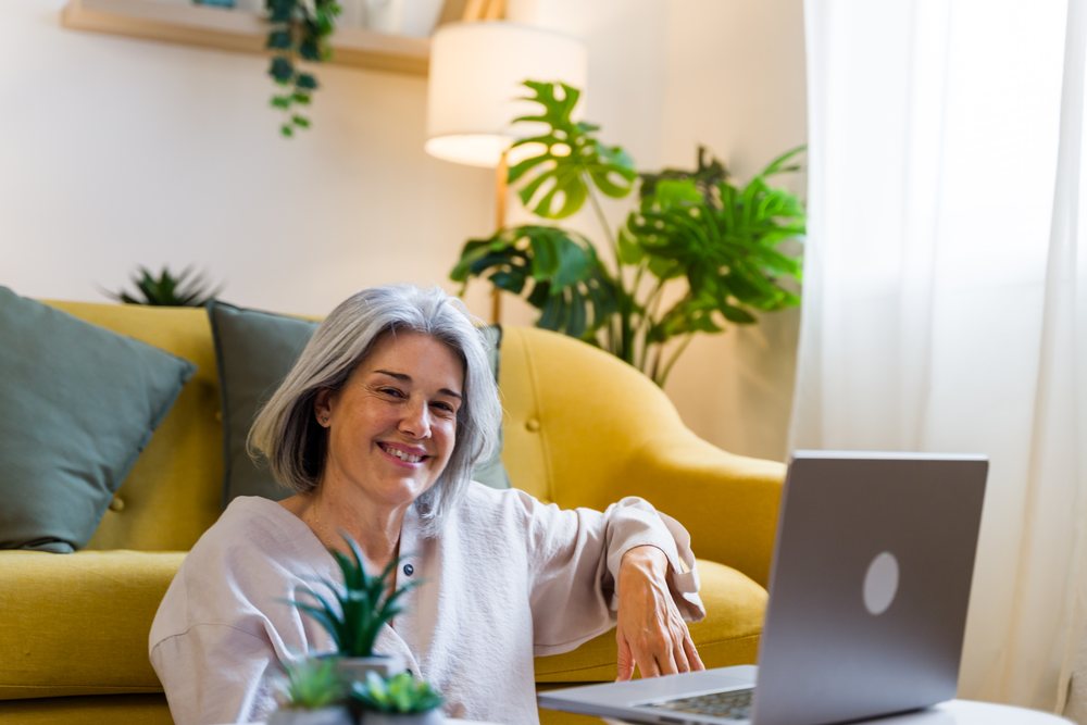 Gray-haired woman smiling while sitting on the floor and using a laptop near a yellow sofa and houseplants in a living room, Spain.