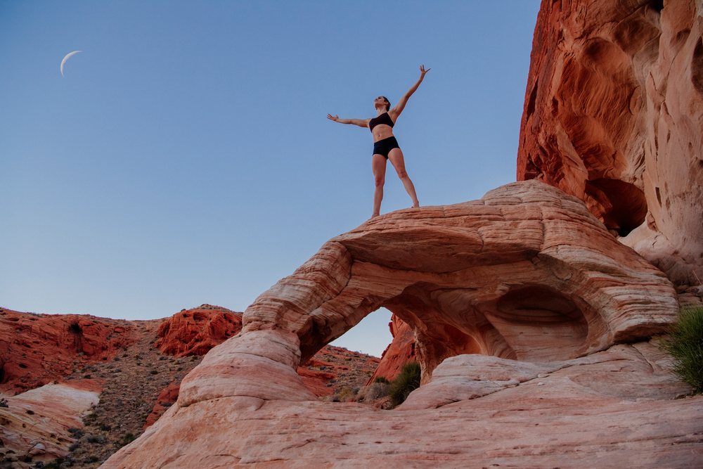 Woman practicing yoga on a red rock formation with a crescent moon visible in the clear desert sky, USA.