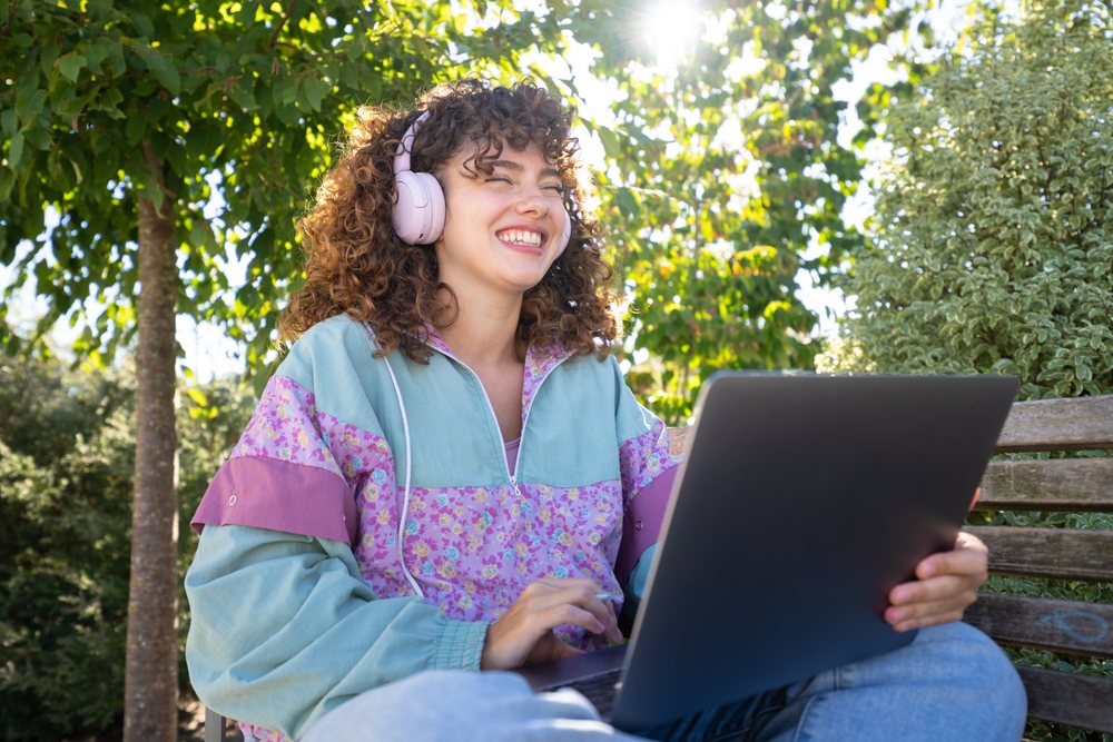 Woman sitting on a bench outdoors using a laptop and wearing headphones, Spain.
