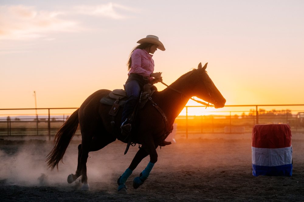 Woman wearing a western hat rides a saddle-equipped horse past a red, white, and blue barrel in a dusty outdoor arena at sunset, Southwest USA.
