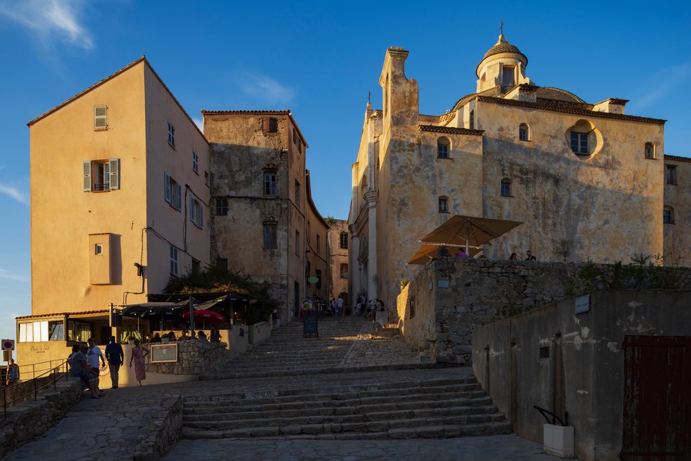Historic Church of St John the Baptist stands atop the Citadel, with people and an outdoor café below during sunset, Travel Photography, Calvi, Corsica, France.