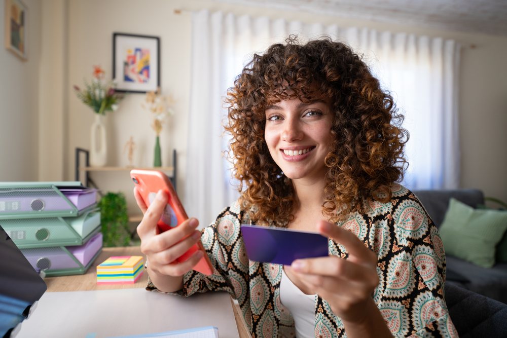 Woman with curly hair smiling while holding a smartphone and credit card at a home office desk, Spain.