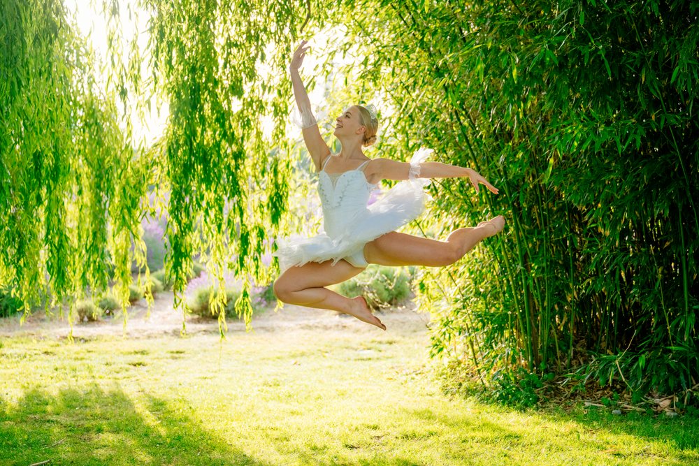 A ballet dancer in a white costume leaps gracefully outdoors under a willow tree, WA, USA.
