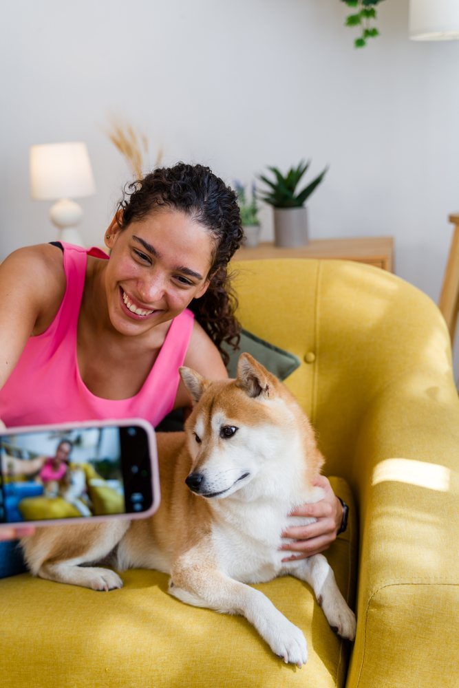 A young woman smiles as she takes a selfie with her dog sitting on a yellow armchair in a living room, Spain.