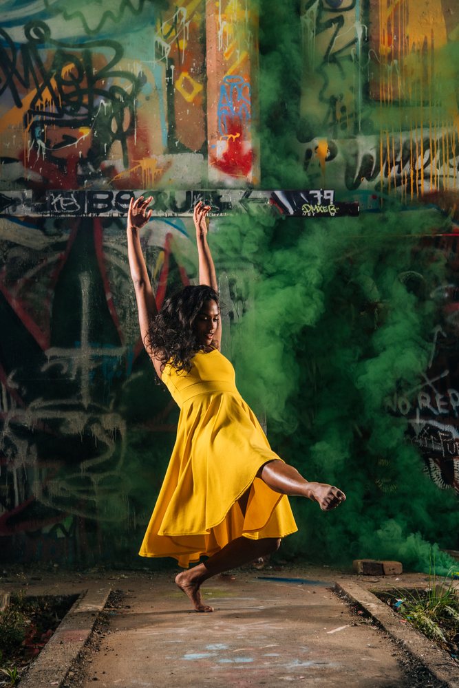 Woman dancing barefoot in a yellow dress with green smoke by a graffiti wall at night outdoors, USA.