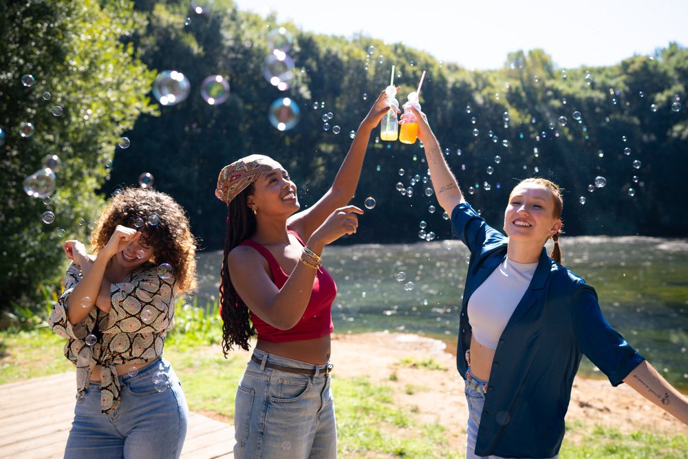 Three women toast beverages surrounded by bubbles outdoors at a sunny lake with trees in the background, Spain.