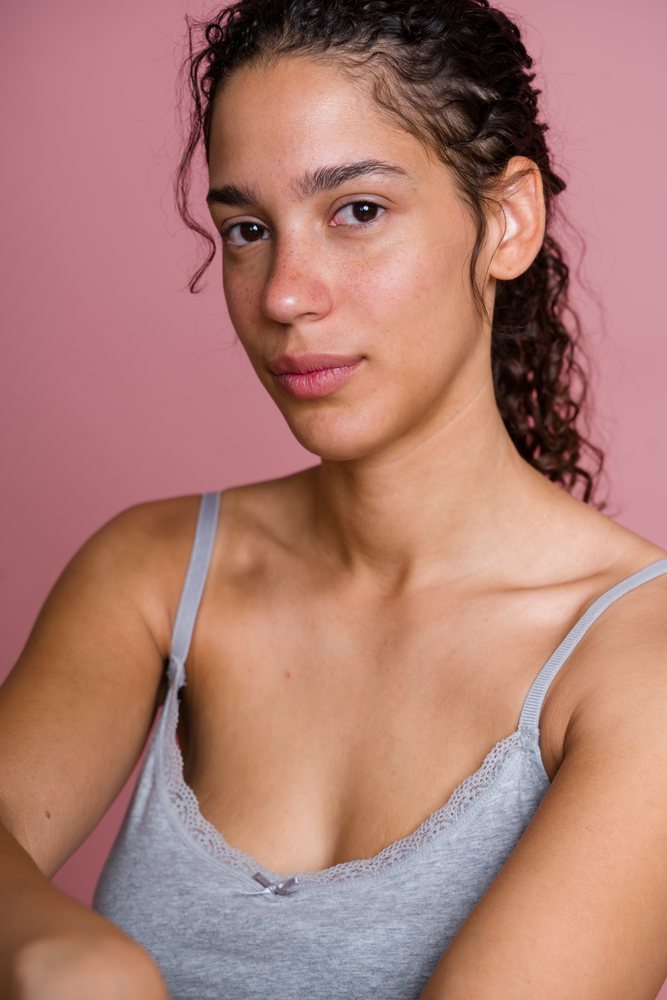 Young woman with curly hair and freckles wearing a gray tank top poses in a pink studio beauty shoot, Spain.