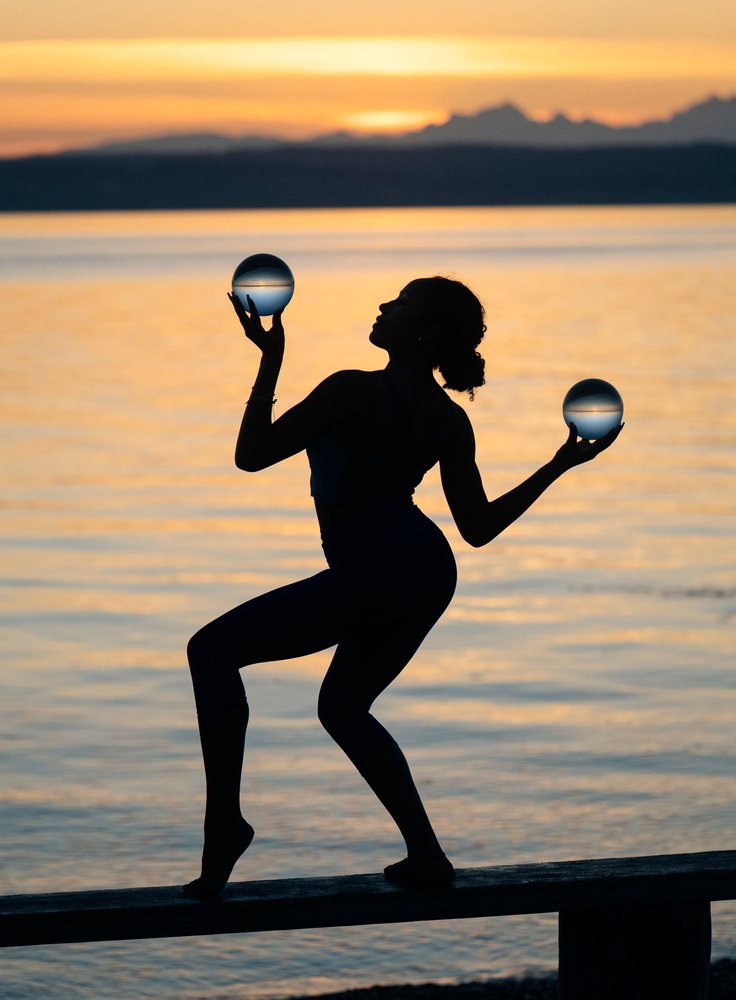 Woman balancing glass spheres while posing in silhouette at sunset beside a lake, yoga, acrobatics, USA.