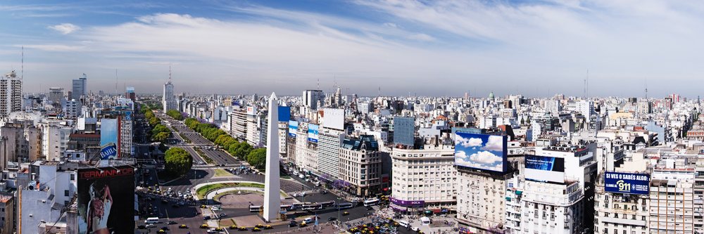 Panoramic view of Buenos Aires cityscape including the Obelisk and busy multi-lane avenue under a blue sky with clouds.