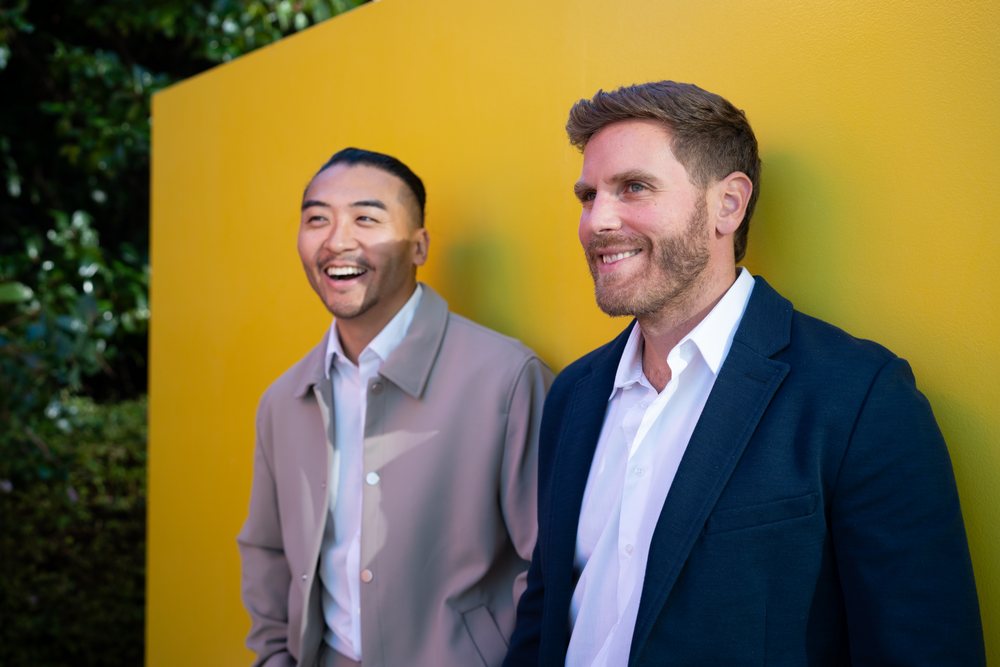 Two men in casual business attire smiling outdoors against a yellow wall, enjoying a cordial moment together in an urban setting, Madrid, Spain.