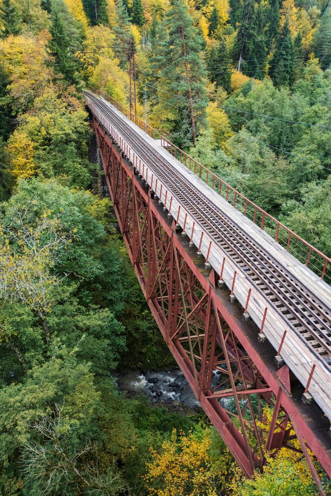 Rust-colored steel truss Railway Bridge with wooden planks over river amid mixed forest with autumn foliage, Tsagveri, Georgia.