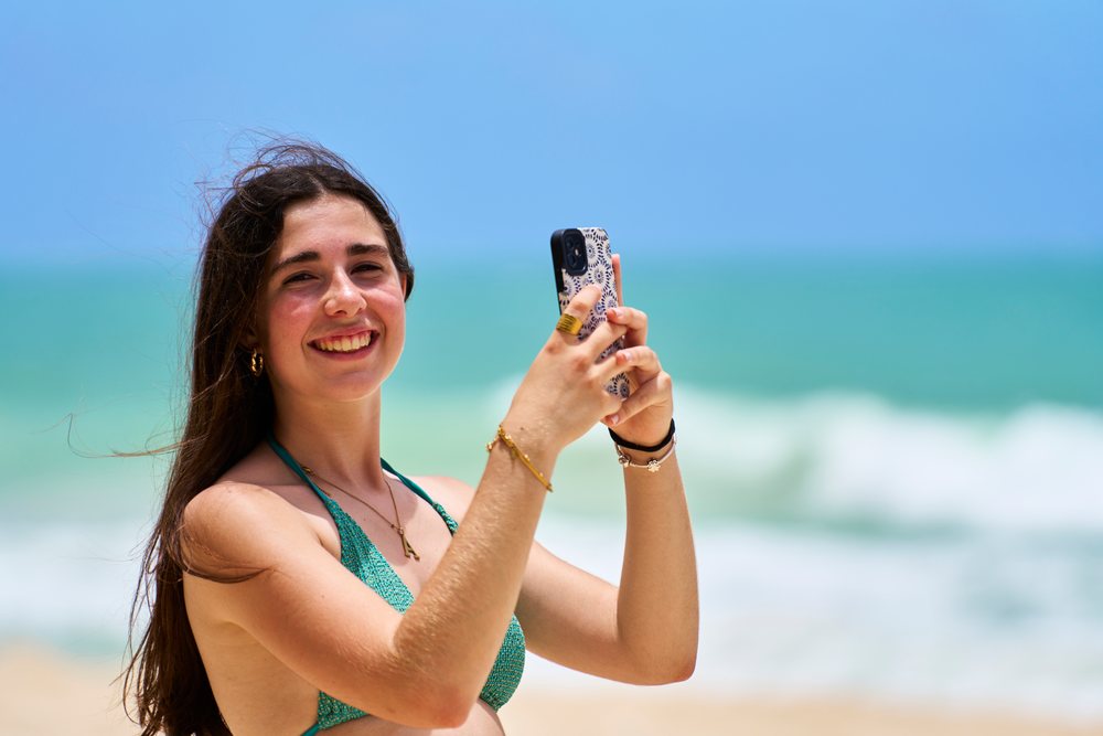 Woman smiling and taking a photo with a smartphone on a sandy beach with ocean waves in the background under clear blue sky, Hawaii, USA.