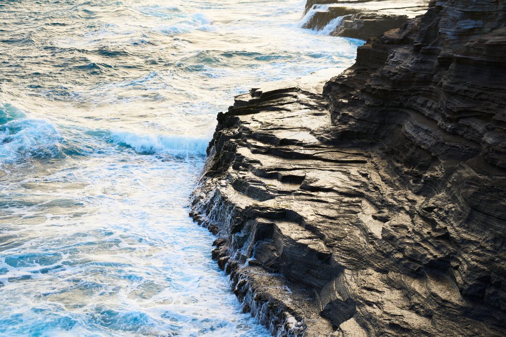 Ocean waves crashing against a rocky cliff on the coast at sunset, Oahu, Hawaii, USA.