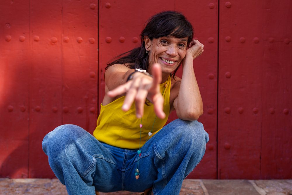 Smiling woman with dark hair wearing a sleeveless yellow top, blue jeans, and a necklace crouches outdoors reaching out in front of a red door, retro, vintage, Málaga, Spain.