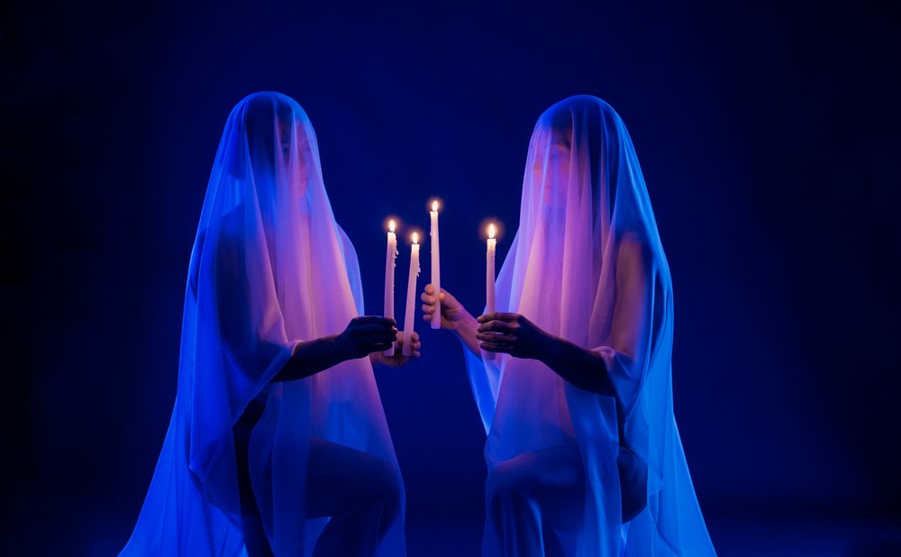 Two people kneeling and holding lit white candles under white translucent veils in blue lighting against a dark background, ghosts, USA.