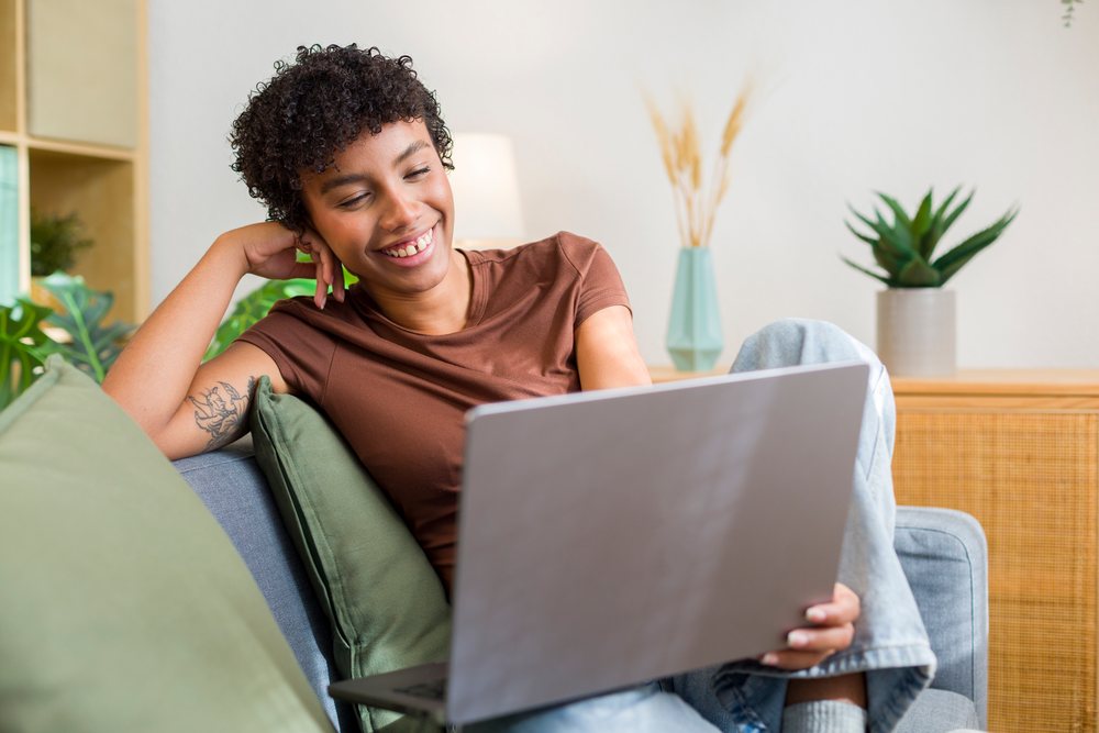 Young woman with curly hair and tattoo sitting on grey couch smiling while using laptop in a decorated home living room.