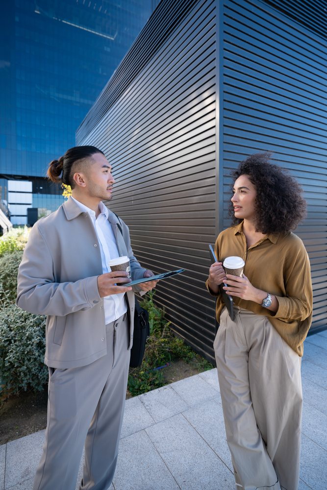 Two professionals stand outdoors by a modern urban building holding coffee cups and tablets during a business discussion, Madrid, Spain.