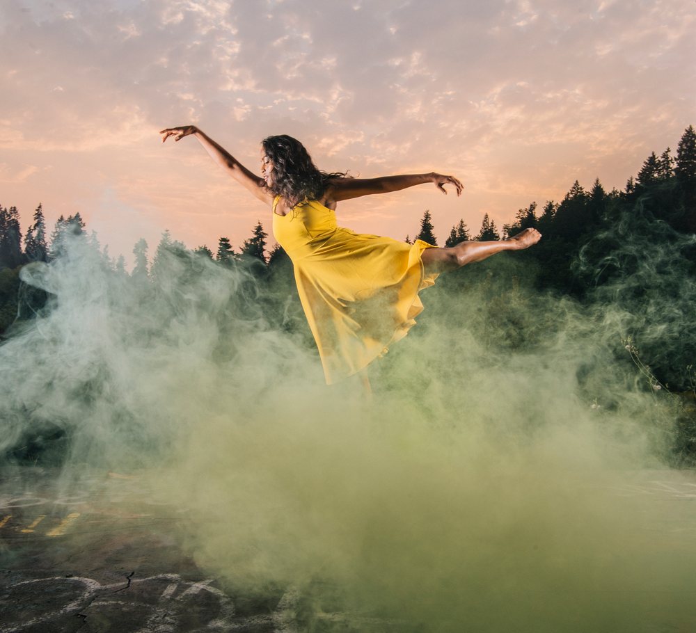 Woman wearing a yellow dress dances gracefully amid smoke outdoors with forest and sunset sky, USA.