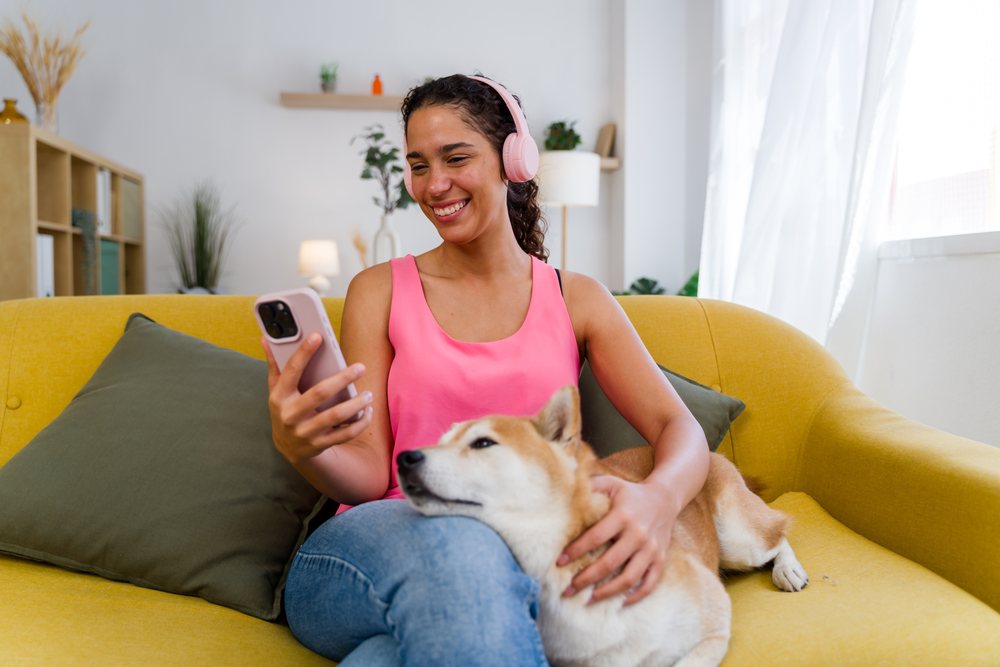 A woman in a pink top wearing headphones listens to music on her smartphone while petting her dog on a yellow couch in a cozy living room, Spain.