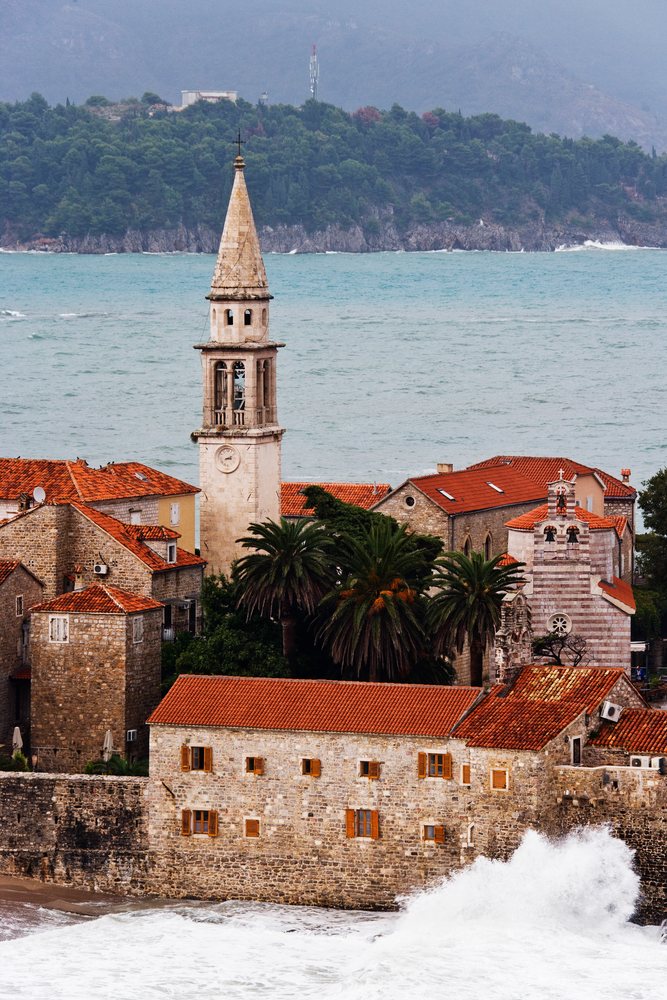 Budva coastal town with historic stone buildings, church bell tower, and waves crashing along the Mediterranean Sea coastline under a mountainous backdrop.