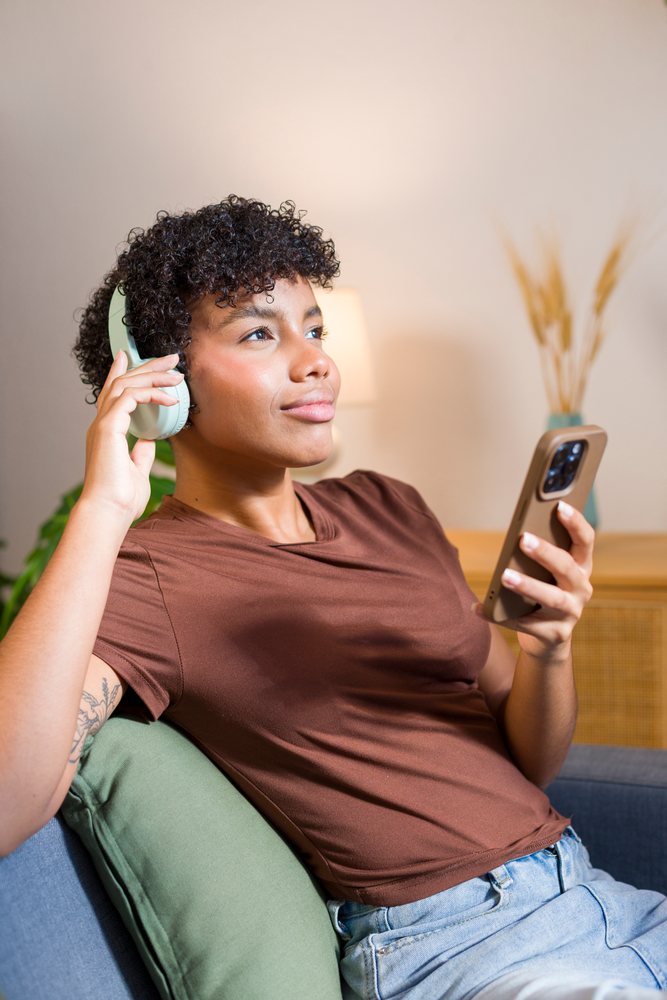 Young woman with curly hair wearing headphones and holding a smartphone while relaxing on a sofa with a green cushion at home.