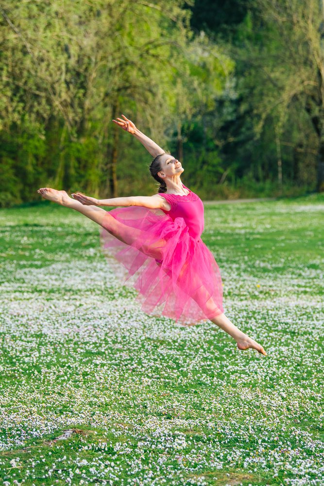 Female ballet dancer in a pink tutu gracefully leaps outdoors in a grassy field with white flowers, USA.