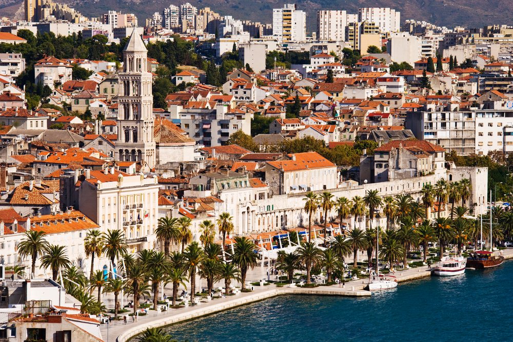 Waterfront view of Split showing historic buildings with red tile roofs, cathedral tower, palm trees along the promenade, and boats docked at the harbor.